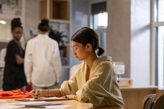Concentrated woman in office setting writing on paper with colleagues in background.