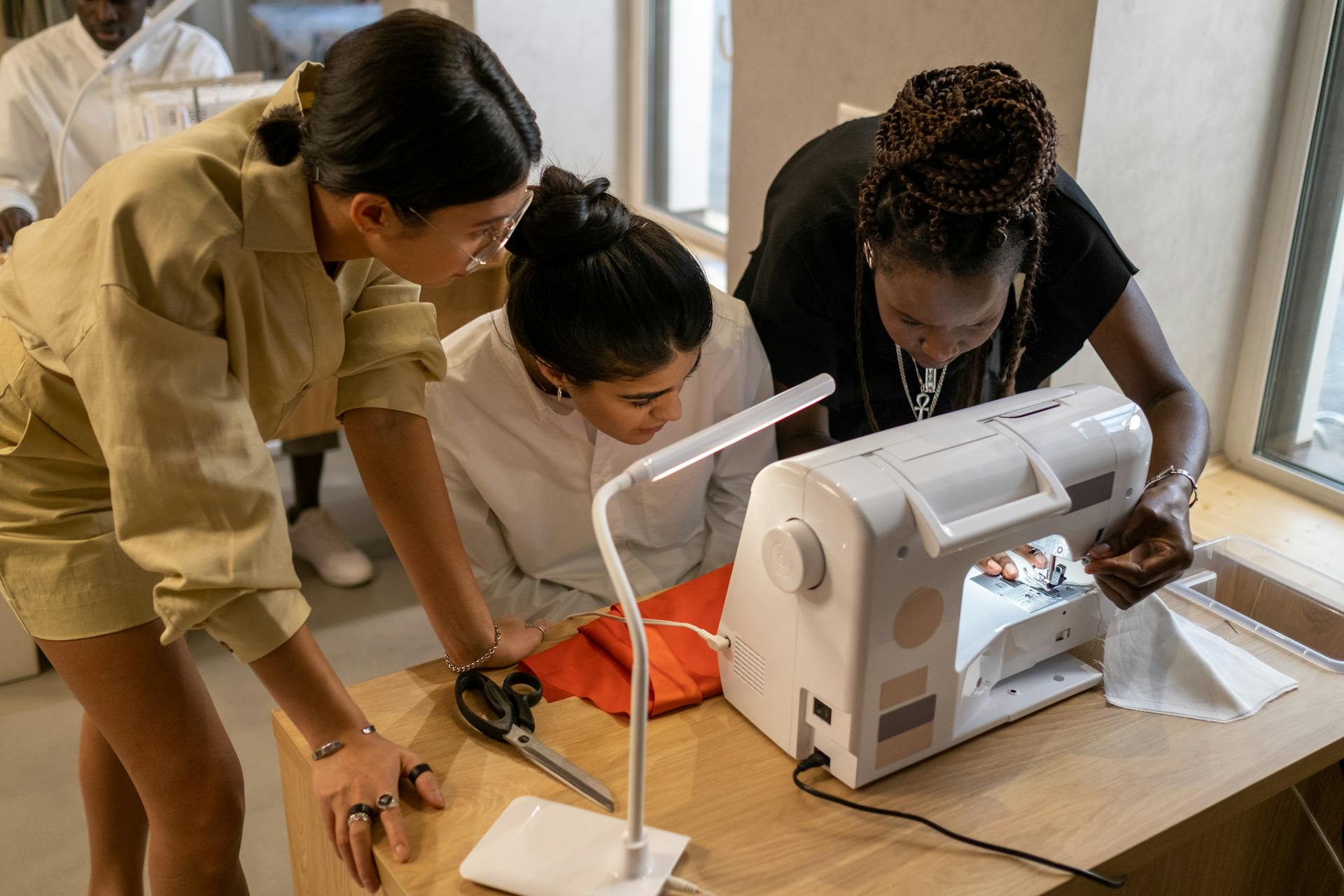 Three diverse fashion designers working together at a sewing machine in a creative studio environment.