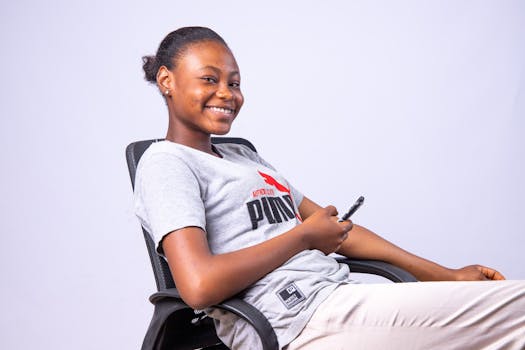 Smiling young woman sitting on a chair indoors, holding a smartphone and relaxing.