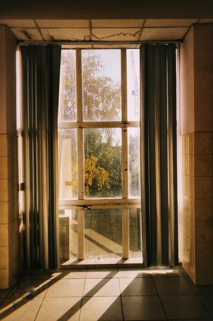 A vertical shot of a window with curtains casting long shadows on the tiled floor, bathed in warm sunlight.