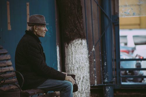 Free A senior man wearing a fedora and black jacket sits on a street bench thoughtfully. Stock Photo