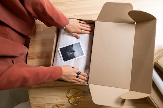 Hands arranging items in a cardboard box with a photo and scissors nearby.