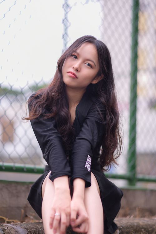 Free Asian girl in black attire sitting pensively by a fence outdoors. Stock Photo