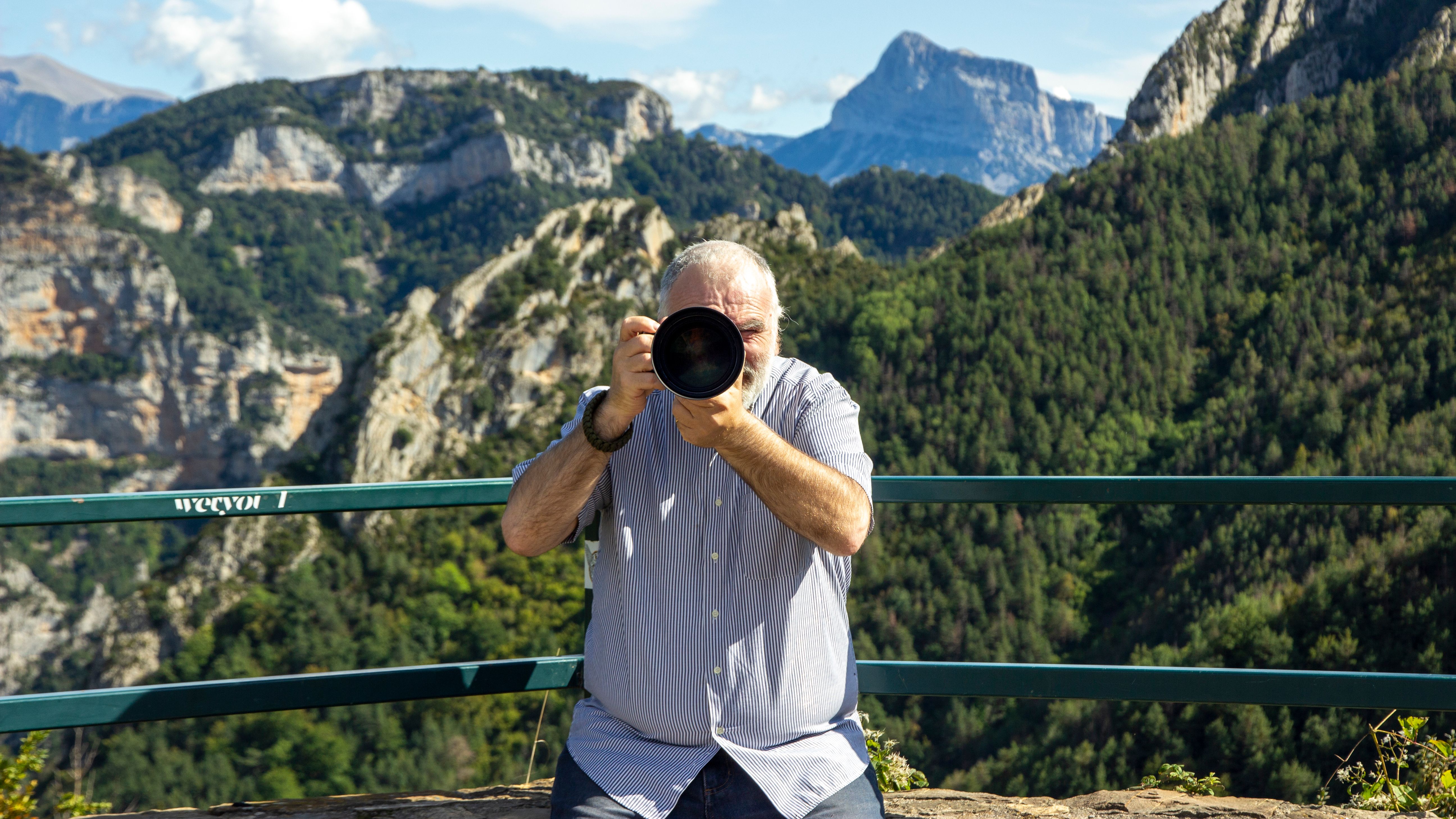 An elderly man taking photos in a scenic mountain landscape.