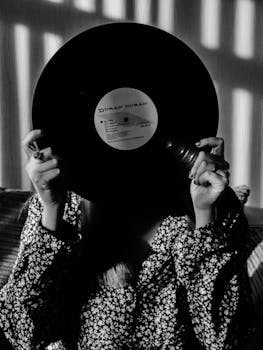 Black and white photo of a person holding a vinyl record, obscuring their face, with striped shadows.