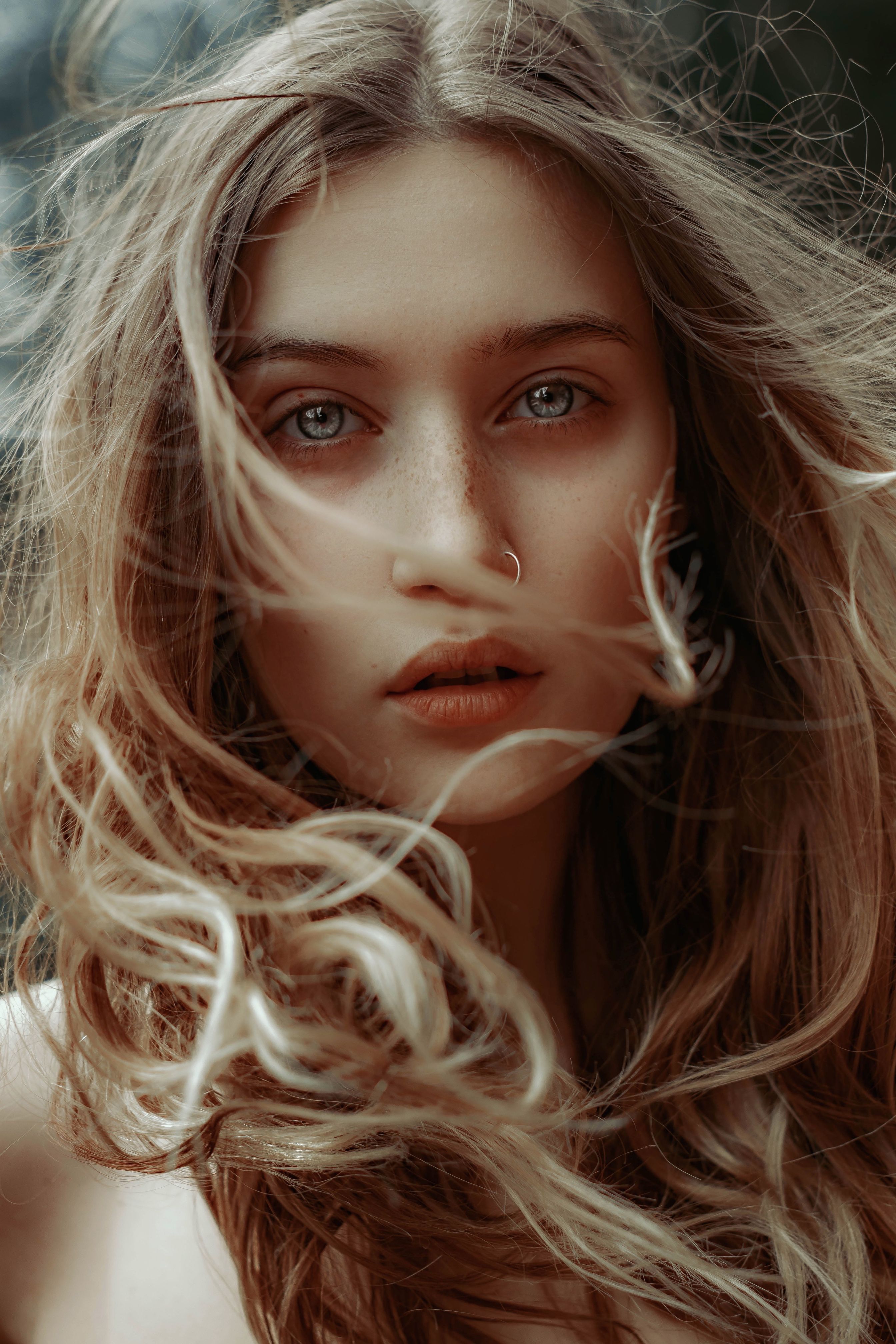 A striking portrait of a young woman with blonde curly hair, featuring a nose piercing.