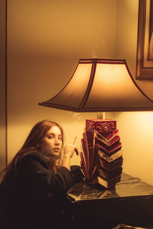 Woman in Black Blazer Sitting Beside Table Lamp Holding a Cigarette 