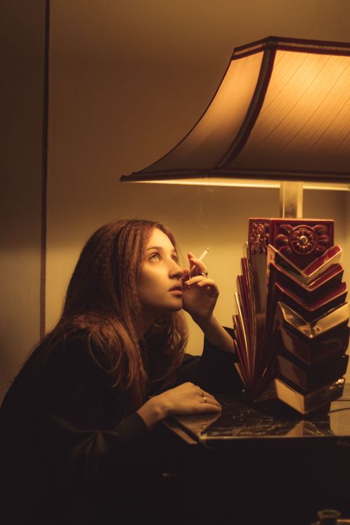 Free A woman in a dark room smokes a cigarette next to a lit table lamp, looking contemplative. Stock Photo