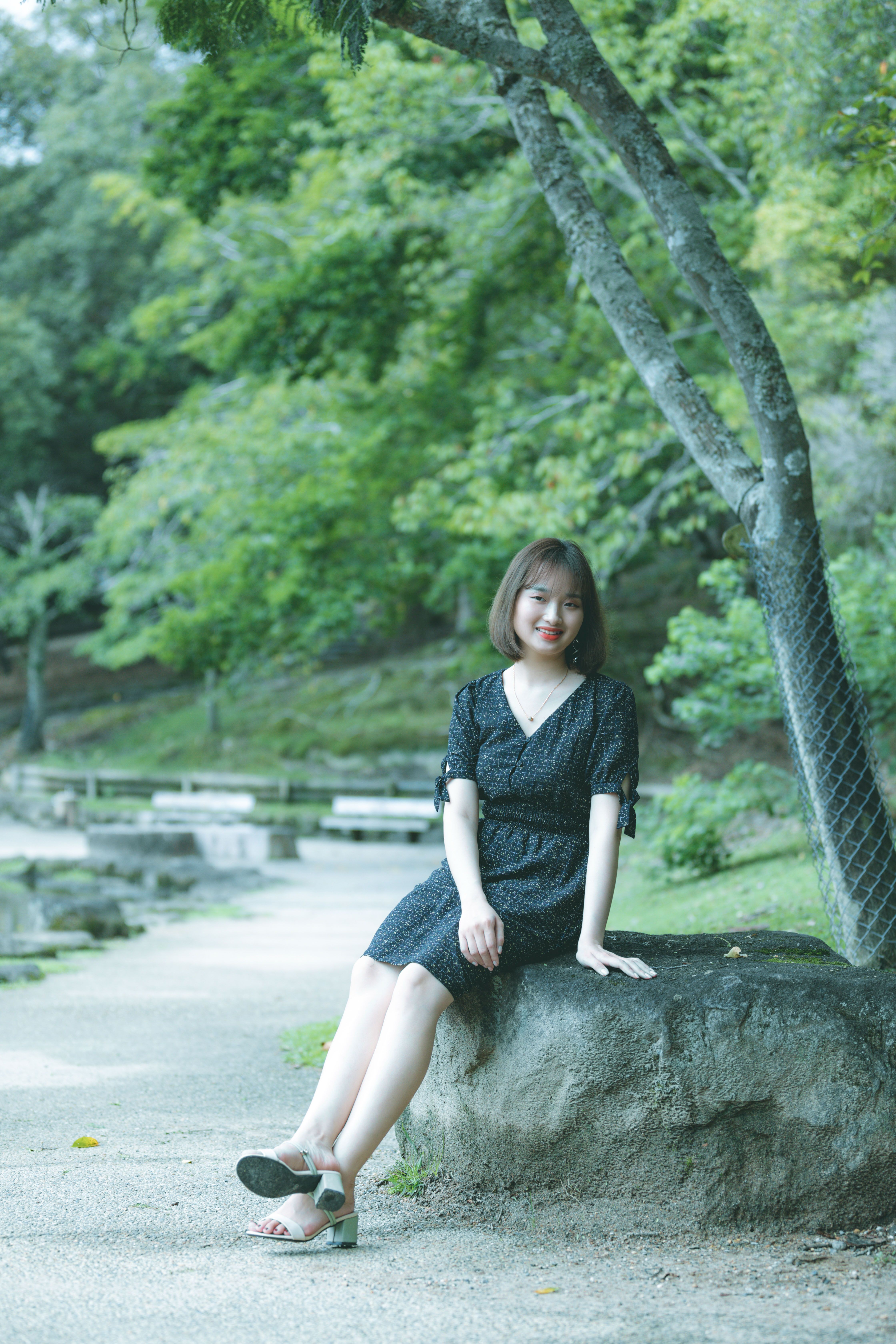 Asian woman in a black dress sitting gracefully on a rock outdoors.