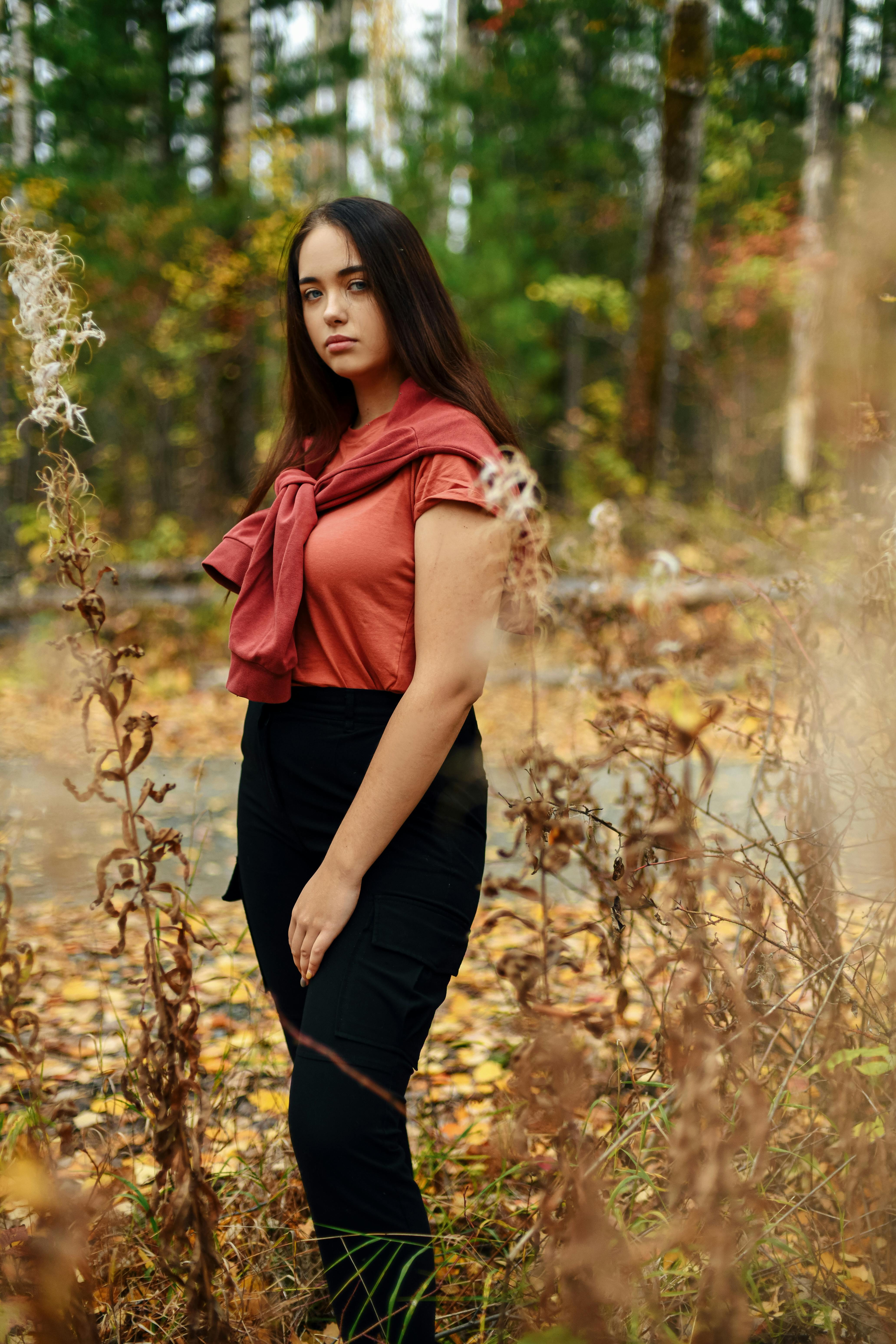 Free Young woman poses in an autumn forest wearing a red t-shirt, surrounded by dried plants. Stock Photo