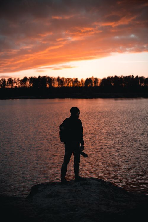 Free A lone explorer silhouetted against a vibrant sunset over a tranquil lake, evoking a sense of adventure. Stock Photo