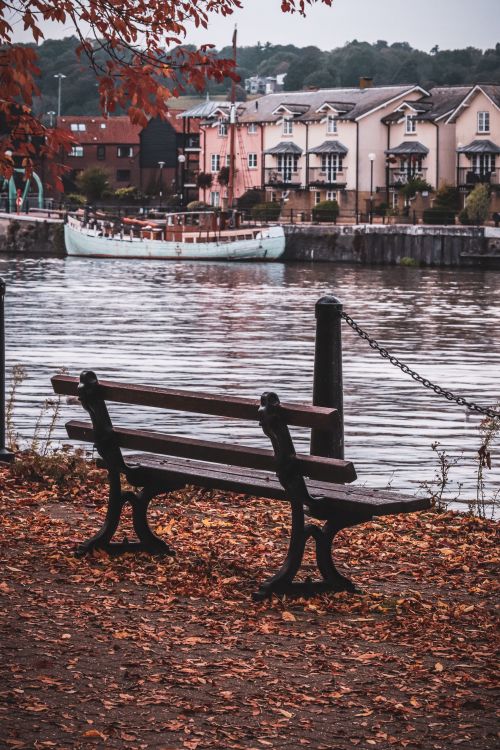 Free Scenic autumn riverbank with empty bench overlooking charming riverside houses. Stock Photo