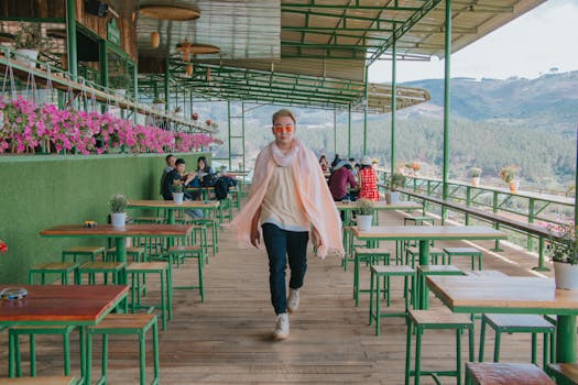A fashionable man walks in a vibrant outdoor café with mountain views and colorful flowers.