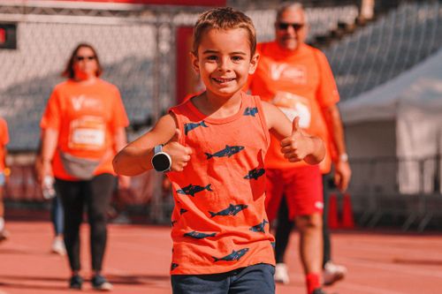 Free Young boy in orange tank top with fish design smiling and giving thumbs up during outdoor event. Stock Photo
