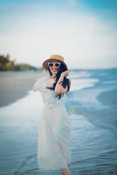 Smiling woman in a summer dress and hat walking on a serene beach.