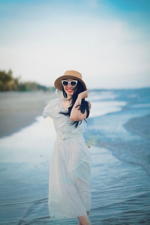 Free Smiling woman in a summer dress and hat walking on a serene beach. Stock Photo