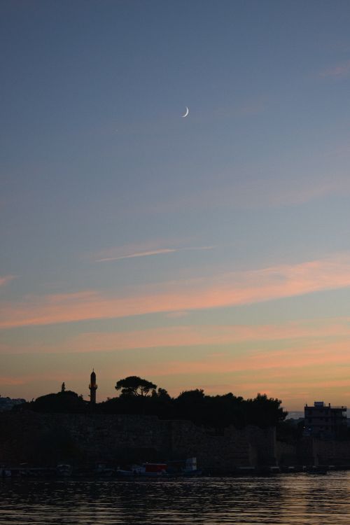 Free A tranquil crescent moon and vibrant sunset sky over silhouetted buildings and trees. Stock Photo