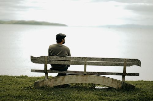 Free A man in a flat cap sits on a wooden bench, enjoying a peaceful lakeside view. Stock Photo
