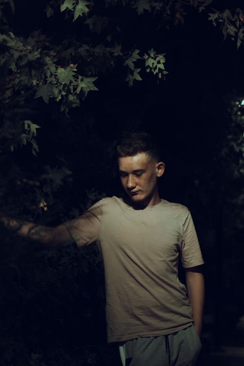 Free A stylish young man poses thoughtfully under tree foliage during a nighttime photoshoot. Stock Photo