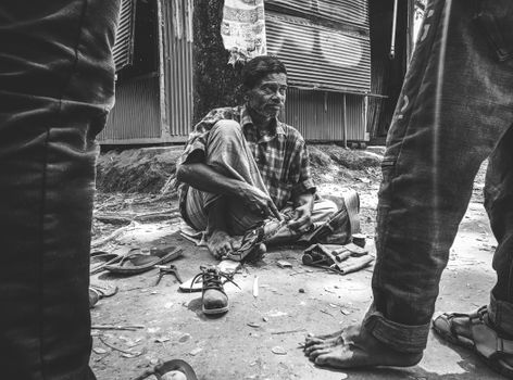 Grayscale image of an adult male cobbler repairing shoes on a busy street.