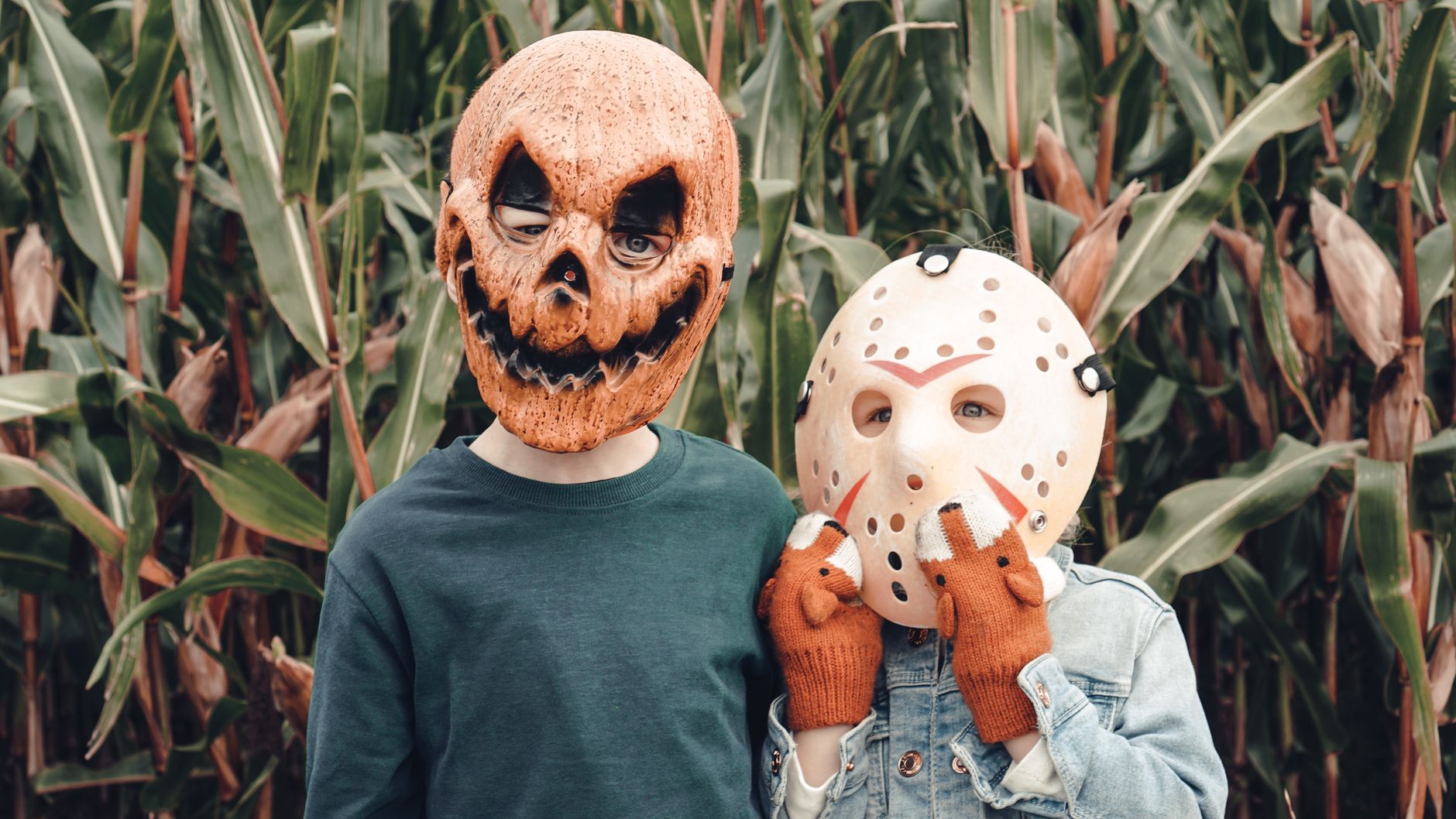 Two children wearing Halloween masks stand in a cornfield, creating a spooky scene.