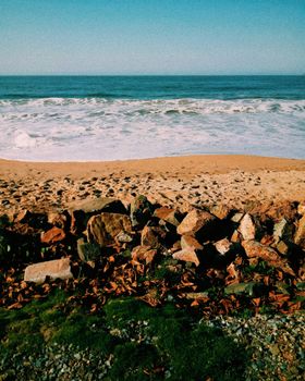 A calm ocean scene with sandy beach, waves, and rocky shoreline under a clear blue sky.