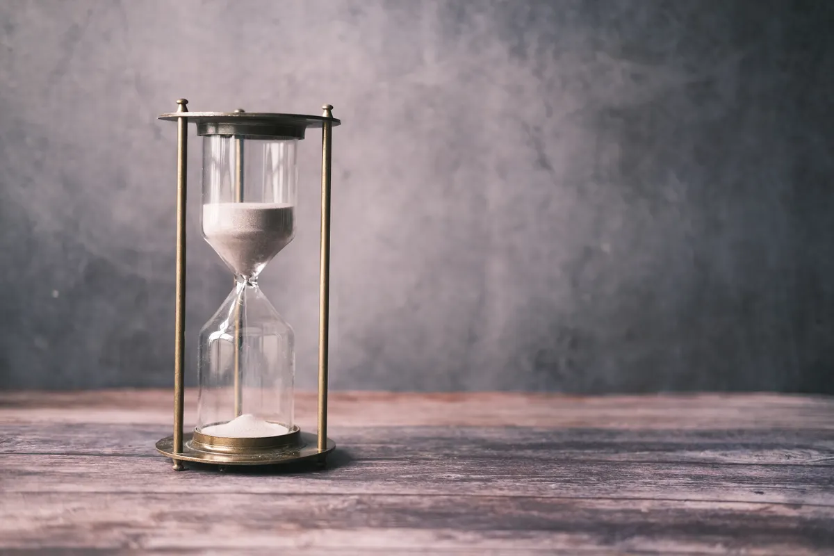 A brass-framed hourglass with pale sand stands on a weathered wooden table against a gray wall.