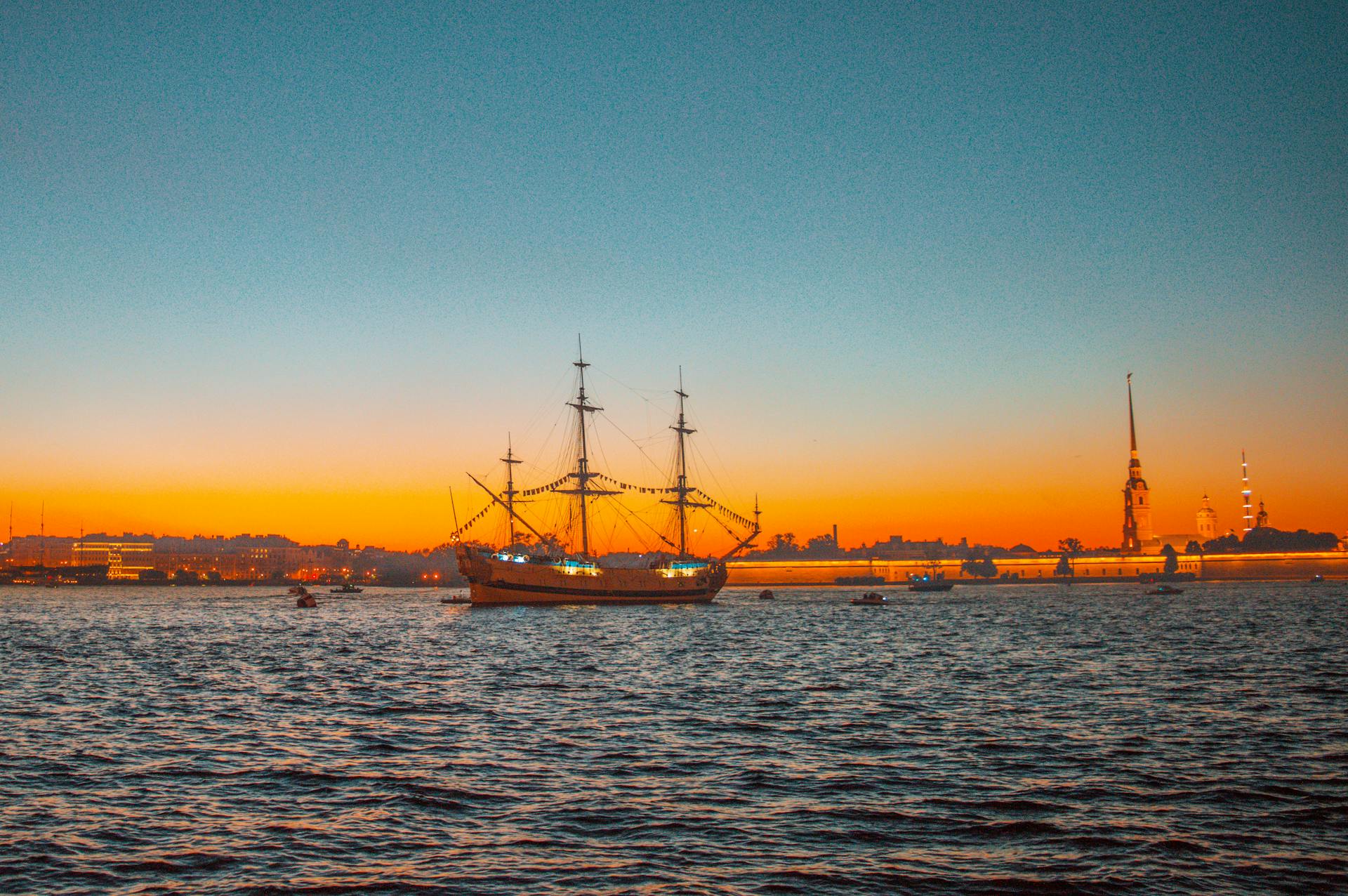 A majestic sailing ship on tranquil waters with a sunset backdrop and historic silhouette.