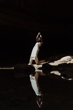 An elegant woman poses in a shadowed cave, reflected in still water below.