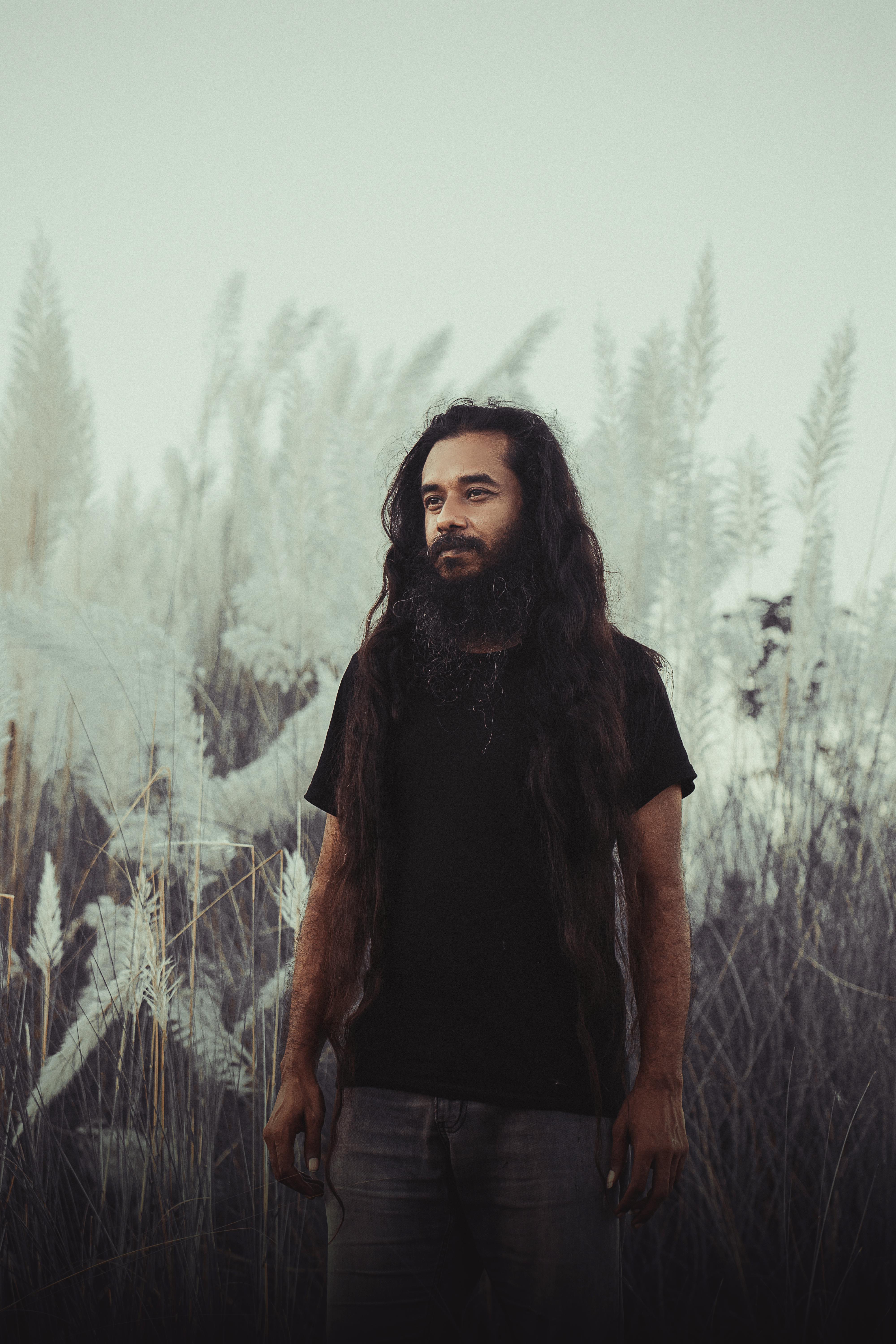 Bearded man with long hair standing in a serene field of tall grasses.