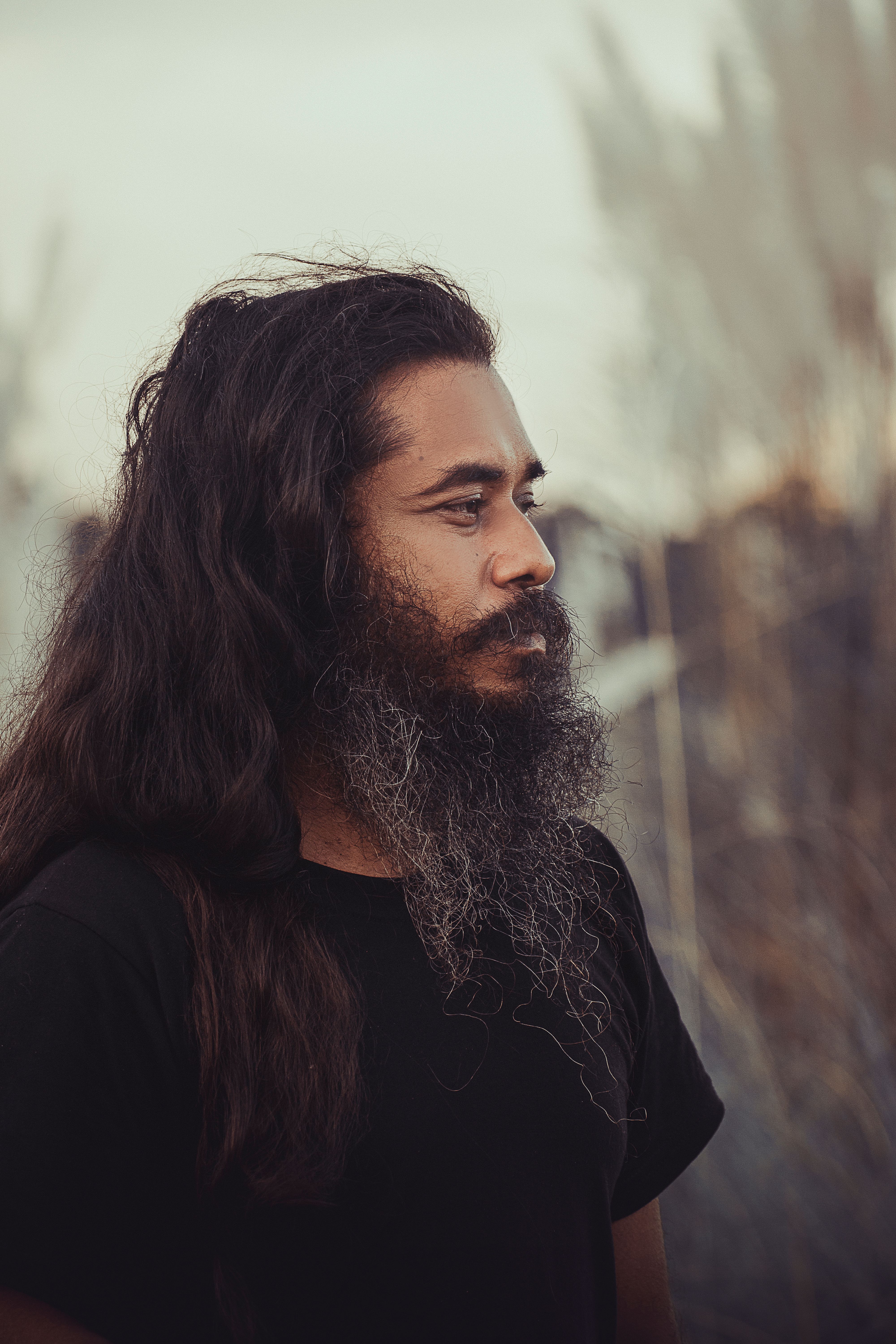 A contemplative man with a long beard and hair, captured outdoors, in a side profile portrait.