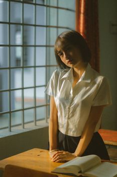 A contemplative Asian woman stands by a classroom window wearing a school uniform.