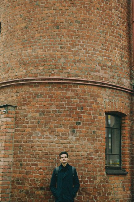 A man in a jacket stands by a red brick wall with a window. Urban setting.