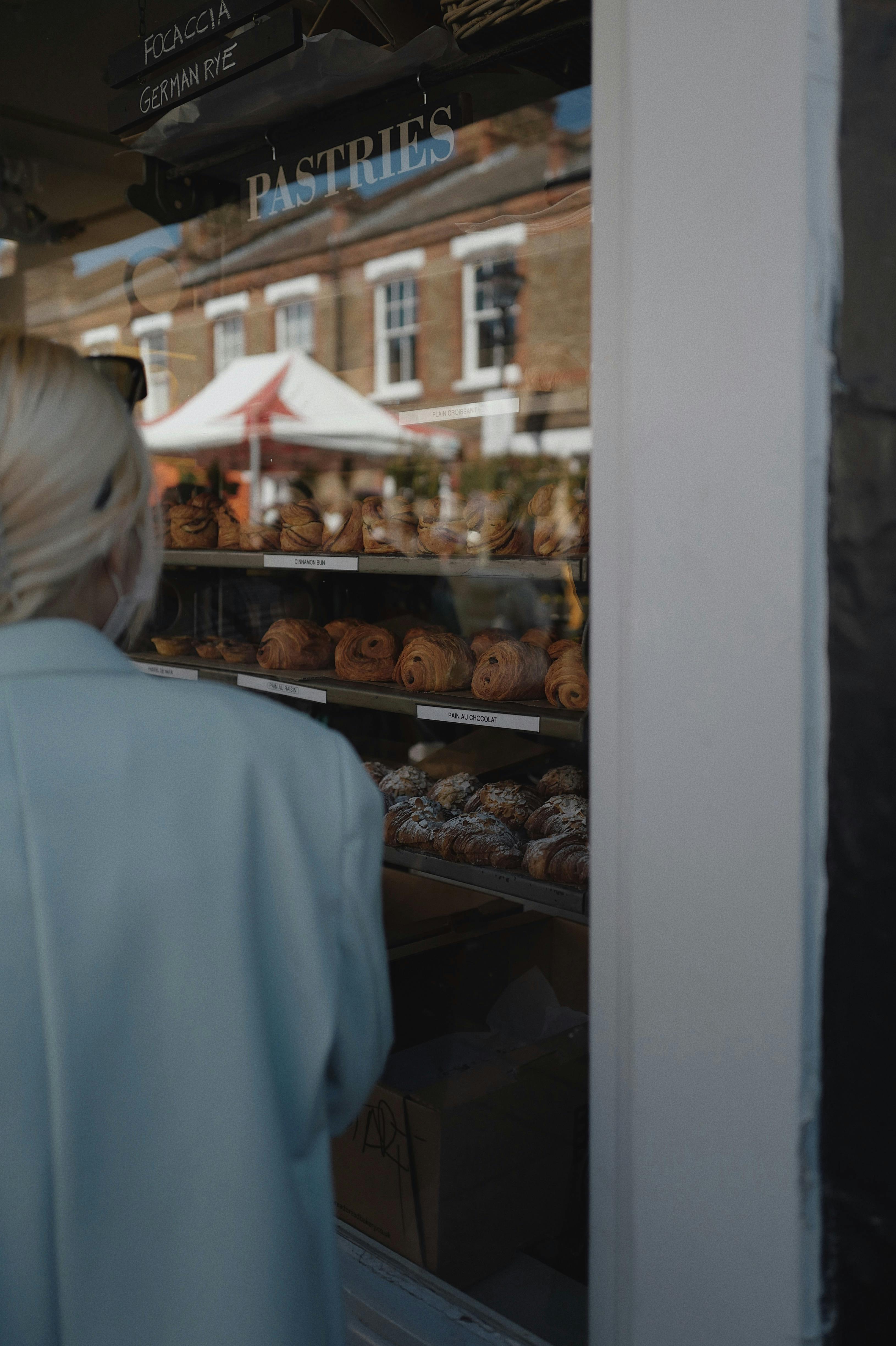 A woman in a coat looking at a variety of pastries through a street shop window display.
