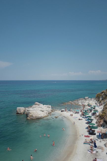 Idyllic beach scene with turquoise waters, rocks, and sunbathers enjoying a summer day.