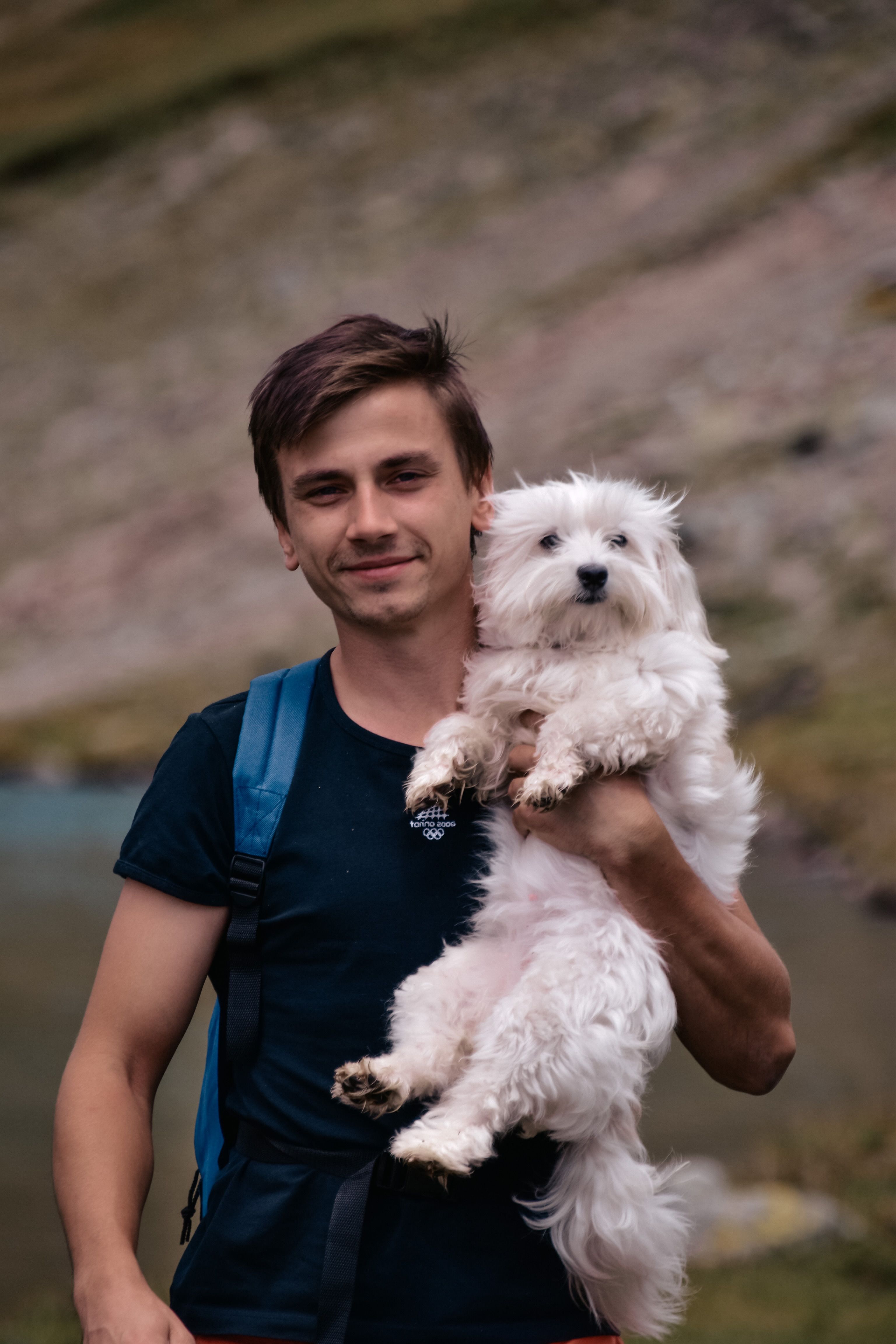A young man in a black shirt holds a white Maltese dog outdoors with a scenic backdrop.