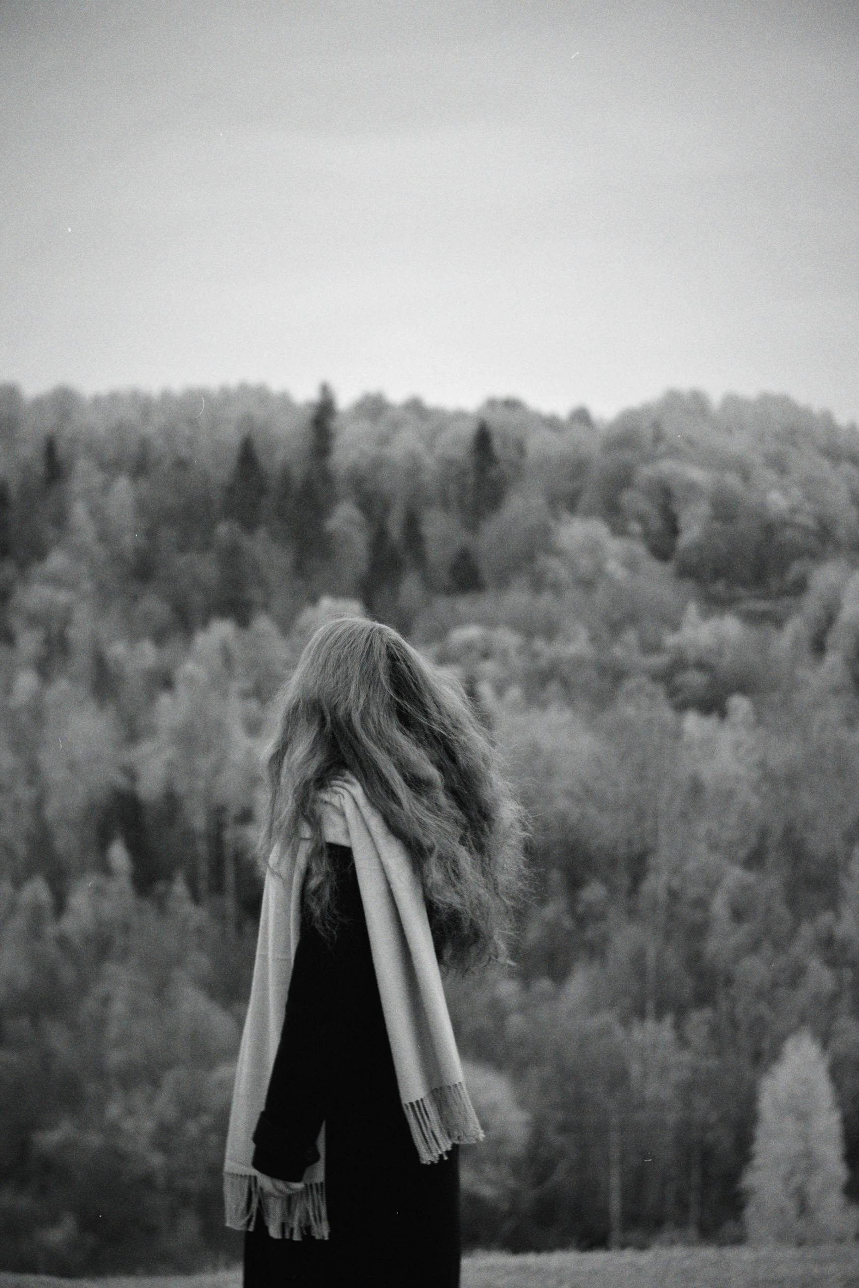 A woman with long hair stands alone, wrapped in a scarf, amidst a serene monochrome forest.