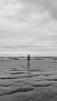 Silhouette of a person walking on Bertioga beach under cloudy skies, captured in black and white.