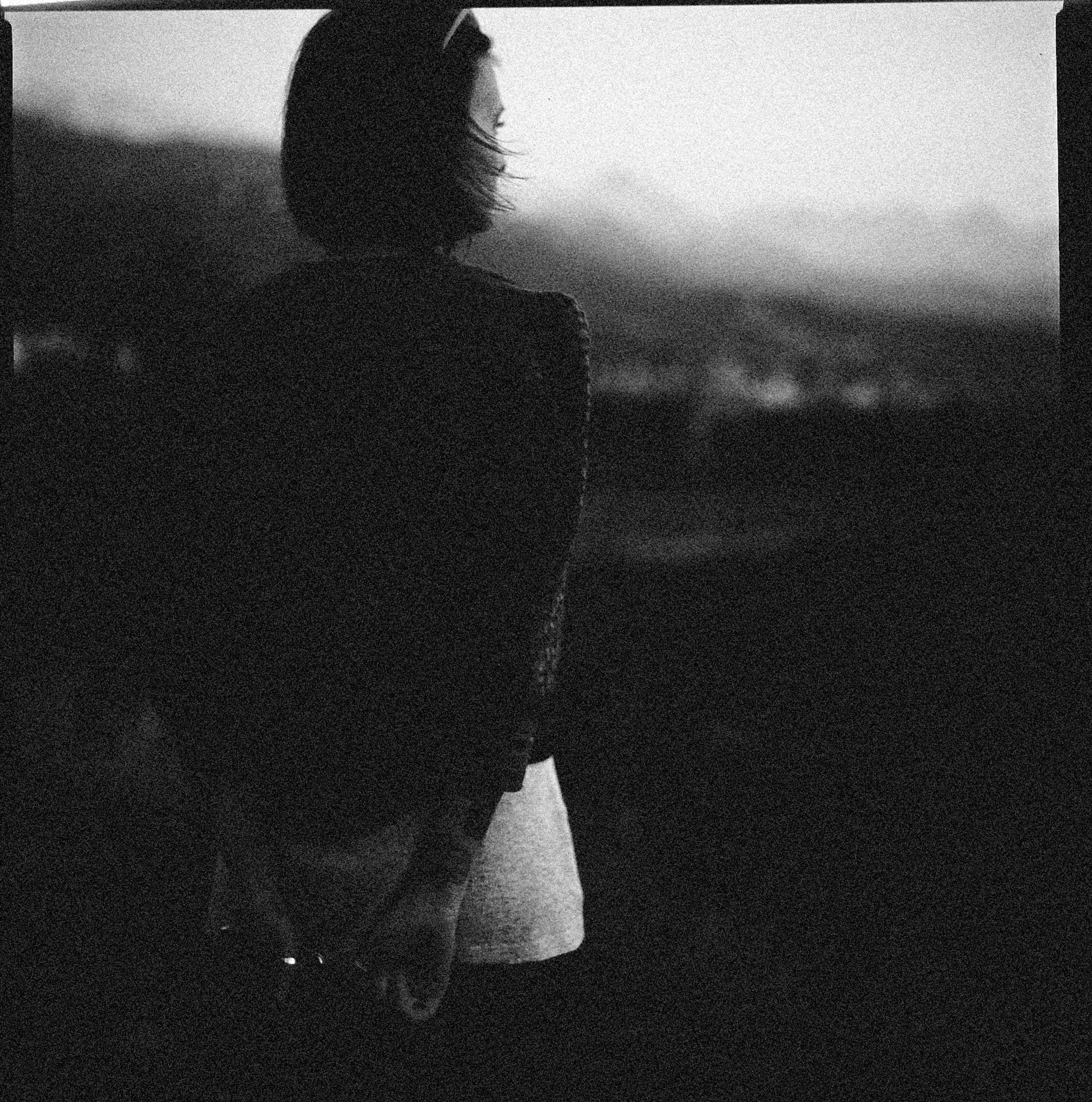 A woman stands alone, gazing into a distant horizon in a grainy, black and white setting.