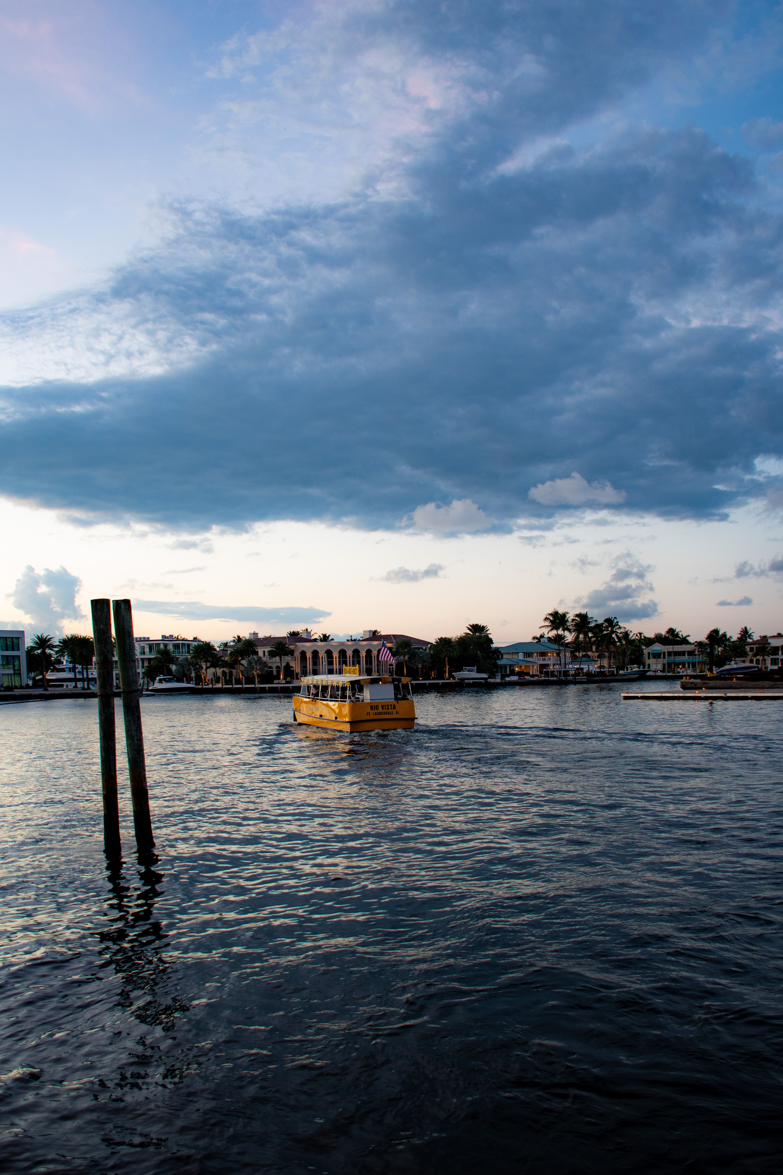 A yellow ferry boat sails on a calm river under a dramatic evening sky.