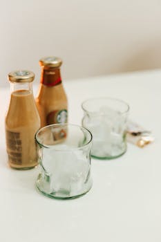 Close-up of bottled iced coffee and empty glasses with ice on a white table.