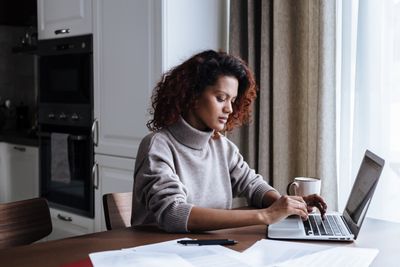 Une femme aux cheveux bouclés travaillant sur un ordinateur portable à une table de cuisine avec des documents