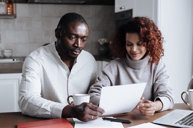 A Black couple sits at a kitchen table reviewing paperwork together, a laptop visible at the edge of the frame; calm engaged register in natural afternoon light.