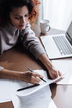 Woman writing on documents while working remotely from home using a laptop.
