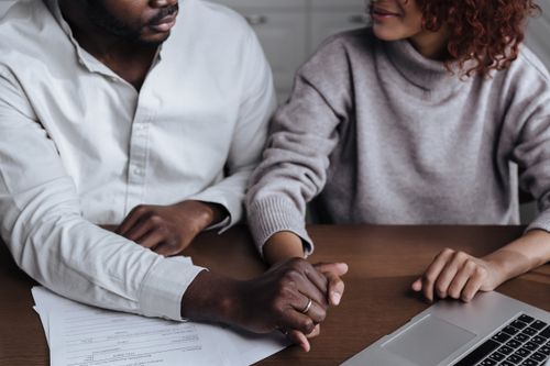 Free A diverse couple holding hands with a laptop and paperwork on the table. Stock Photo