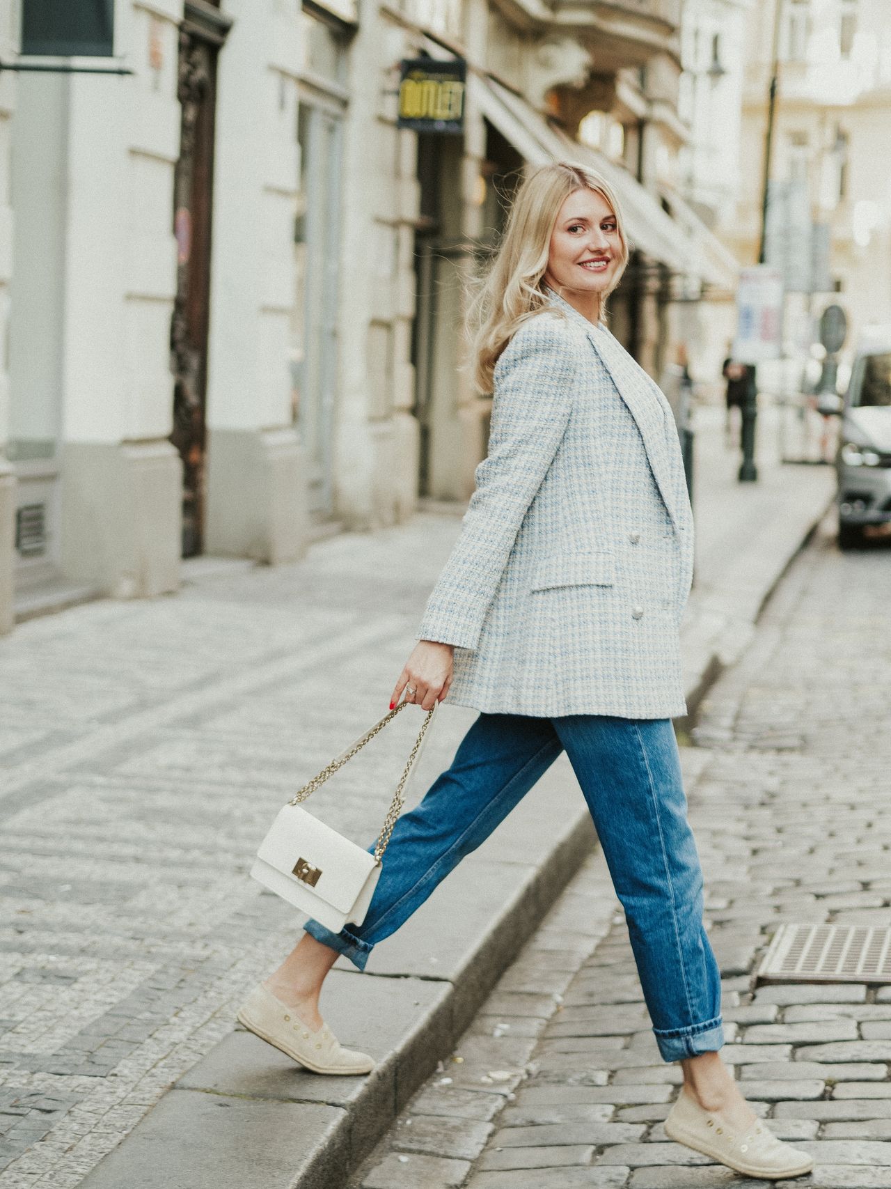 Woman in a light blue tweed blazer, cuffed dark jeans, beige flats, and a white chain-strap bag walking on a cobblestone street.