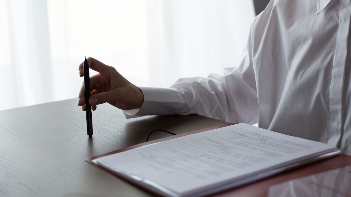 A woman examines documents, holding a pen over a table in a bright indoor setting.