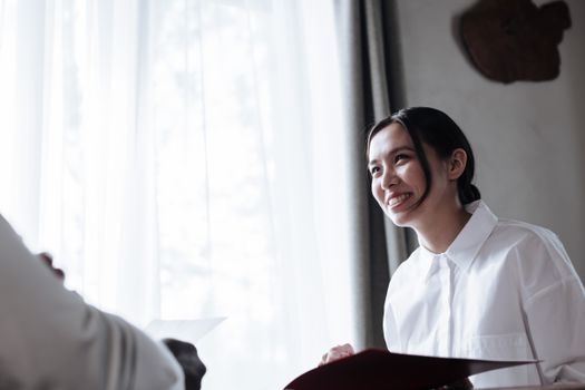 A smiling woman in white shirt engaged in conversation indoors by a window.