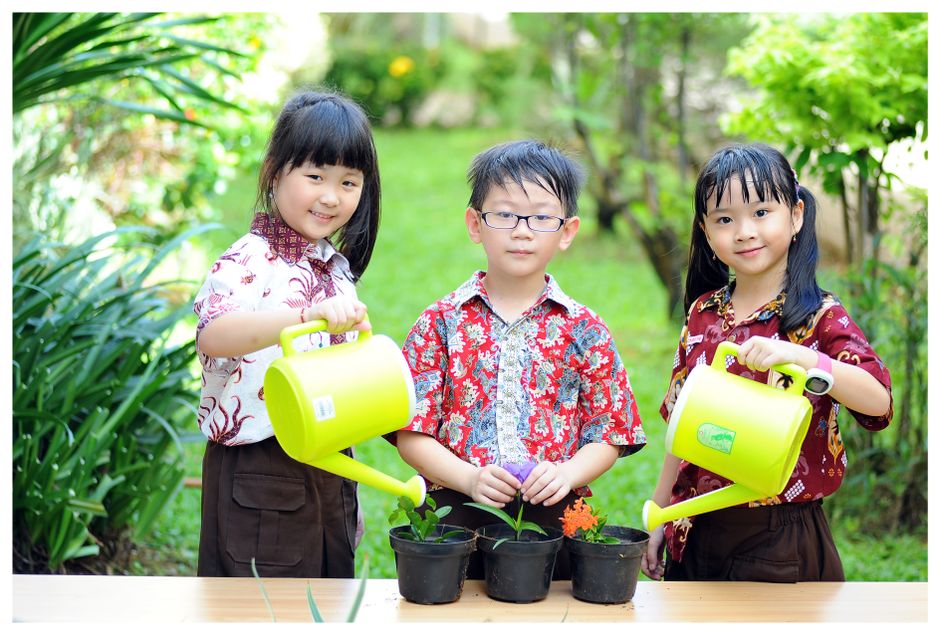 Three children watering plants in a lush Jakarta garden, smiling with colorful watering cans.