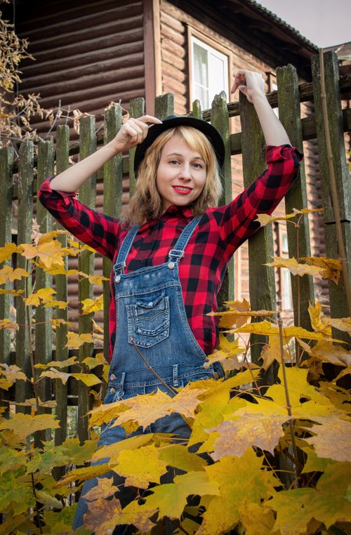 Free A joyful woman in denim overalls and a plaid shirt poses playfully with autumn leaves. Stock Photo